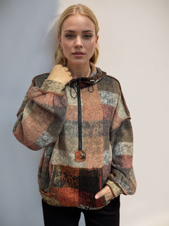 This image shows a young woman posing against a neutral grey background. She is wearing a stylish, oversized jacket featuring a patchwork design with muted colors like grey, brown, and rust. The jacket has a high collar with a black zipper and is made of a cozy, textured fabric. Her blonde hair is styled back, and she has a subtle makeup look, highlighting her natural beauty.